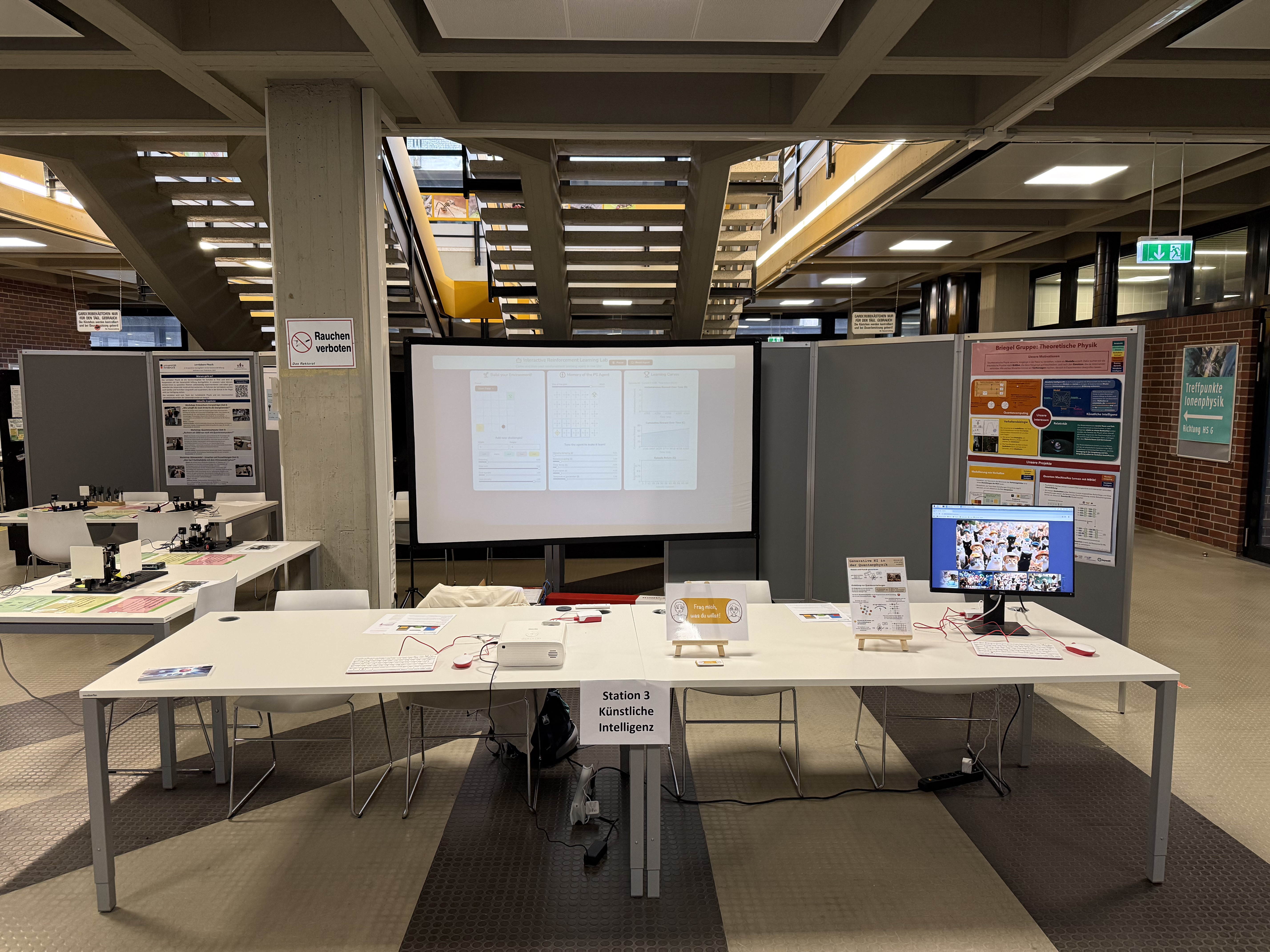 Layout of the booth. The projector screen displays our interactive reinforcement learning demo, with keyboard and mouse near the projector for interactivity, and the group poster is positioned to the right of the projector screen. The "ask me anything" station is to the right of the projector and the circuit generation game is to the right of that.